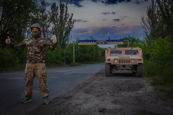 A soldier in camouflage stands by a military vehicle on a rural road at dusk.