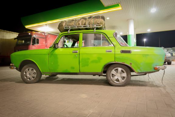 A vintage green car parked at a gas station, with luggage on the roof.