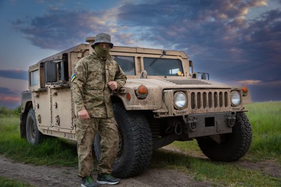 A soldier in camouflage stands beside a beige Humvee on a grassy field under a cloudy sky.