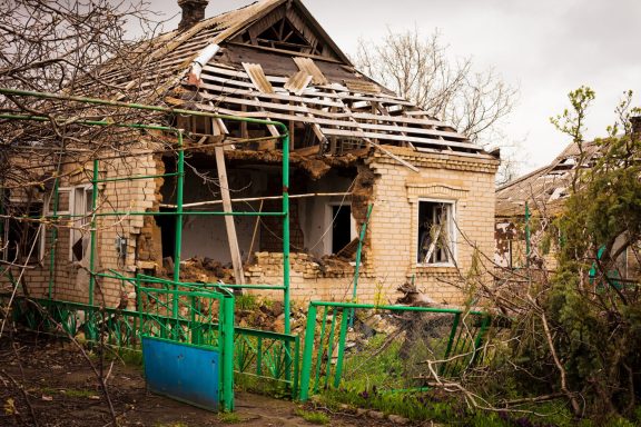Damaged house with a collapsed roof and broken windows, surrounded by greenery.